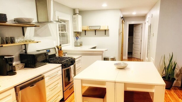 A compact kitchen with white cabinetry, a dishwasher, a stove, and a small dining area with stools.