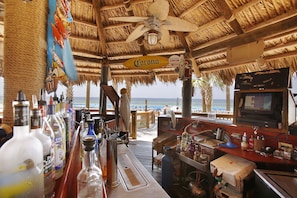 Um bar de praia com telhado de palha, um letreiro da Corona e vista para o oceano.