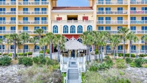 Un hotel de varios pisos con una entrada central, rodeado de palmeras y un camino bordeado de playa que conduce a la entrada.