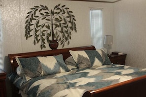 Een slaapkamer met een houten bed, twee kussens, een decoratieve wandversiering, een nachtkastje en een lamp.