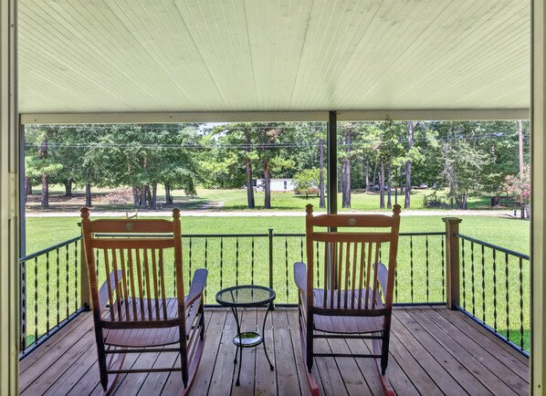Eine überdachte Veranda mit zwei Holzstühlen und einem kleinen Tisch, mit Blick auf eine Rasenfläche mit Bäumen und ein Gebäude in der Ferne.