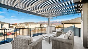 A covered outdoor patio with wicker furniture, a table, and a view of a residential area with mountains in the background. - The Legacy Retreat Near National Parks! (Hurricane)