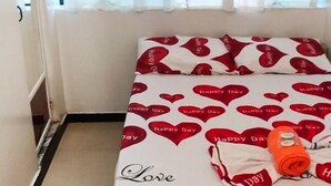A bedroom with a bed covered in a white and red heart-patterned bedspread, a window with white curtains, and a small orange item on the bed. - Galo Vela (Santo Domingo de los Colorados)