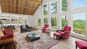 A spacious living area with a high ceiling, large windows, and a view of the outdoors. - NEWLY RENOVATED! Lake George Mtn Retreat: Hot tub, BBQ, Firepit, 2-tiered Deck! (Lake George)