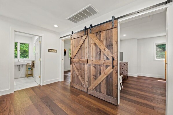 A room with a sliding barn door, hardwood flooring, a white wall, and a window with a view of greenery. - NEWLY RENOVATED! Lake George Mtn Retreat: Hot tub, BBQ, Firepit, 2-tiered Deck! (Lake George)