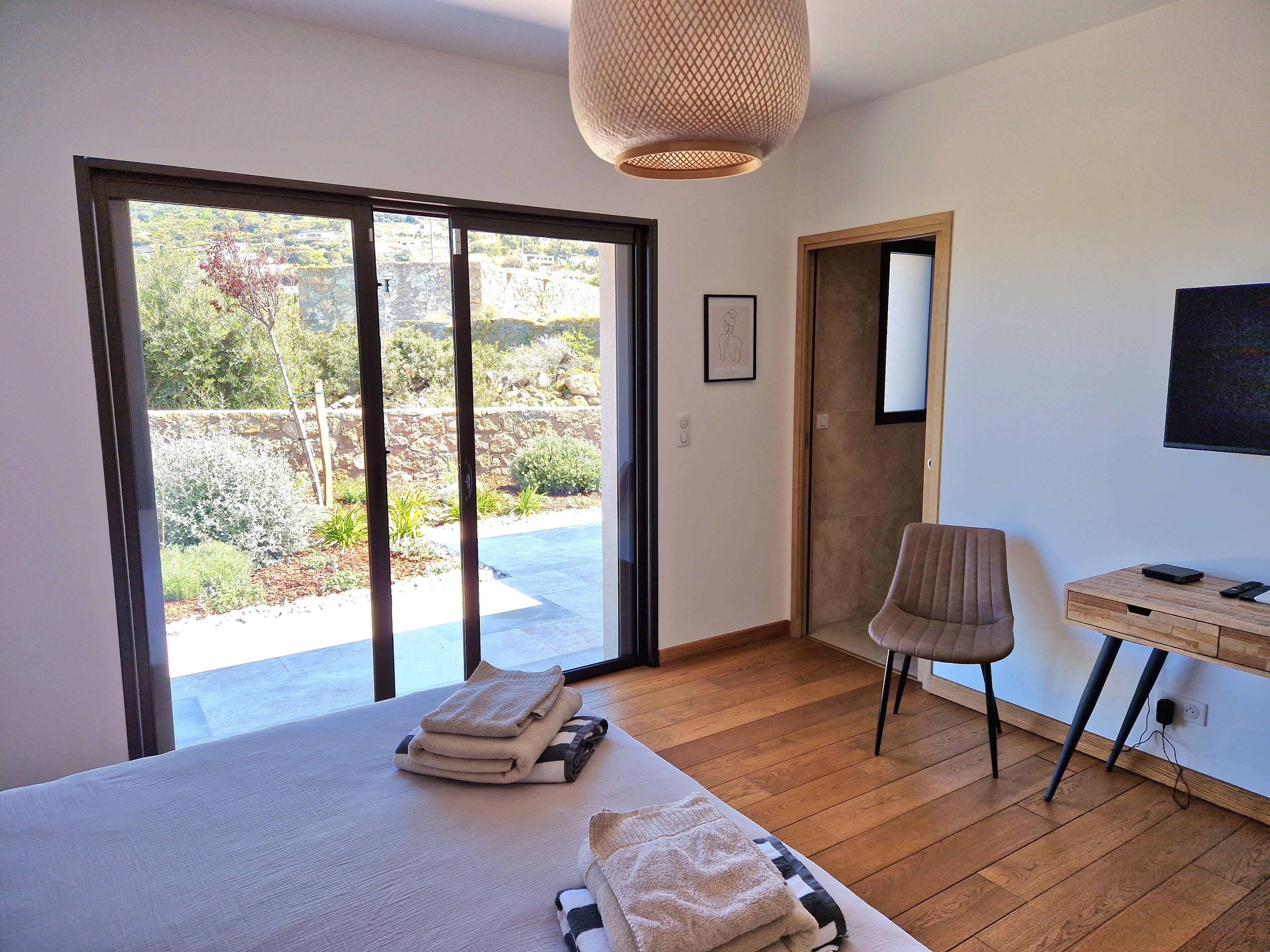 A bedroom with a large bed, a desk, a chair, and a view of the outdoors.
