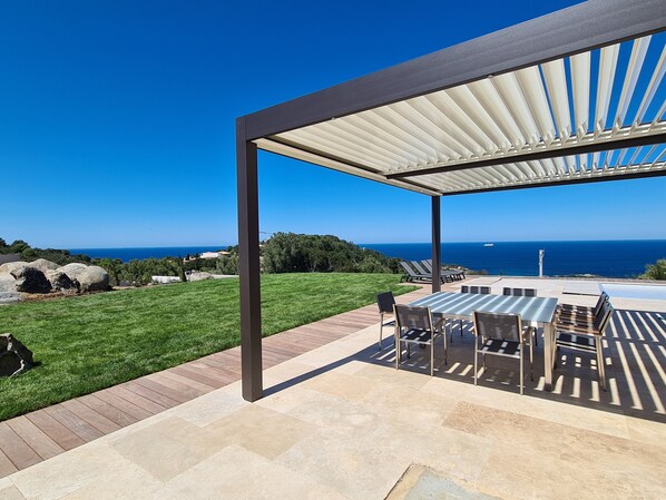 Une terrasse extérieure couverte avec vue sur l’océan, une pelouse verdoyante et un chemin en pierre.