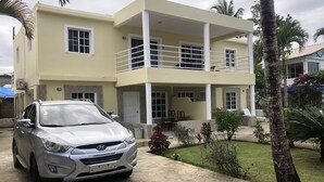 A two-story house with a balcony, a car parked in front, and a well-maintained garden with palm trees and flowers.