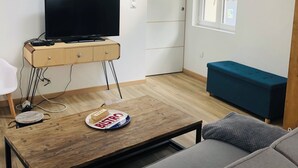 Una sala de estar moderna con un televisor de pantalla plana sobre un mueble de madera, un sofá azul, una mesita con un plato de galletas y una ventana con persianas.