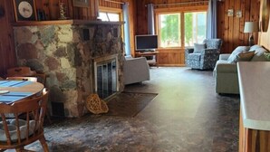 Een gezellig blokhutinterieur met een stenen open haard, houten wanden, een eethoek en een zithoek met een televisie.