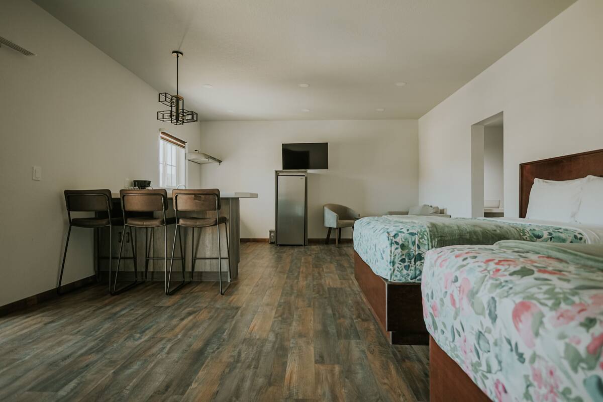 Ein Hotelzimmer mit Holzboden, einem Bett mit Blumenmuster-Bettwäsche, einem Schreibtisch mit Stühlen, einem Kühlschrank und einem an der Wand befestigten Fernseher.