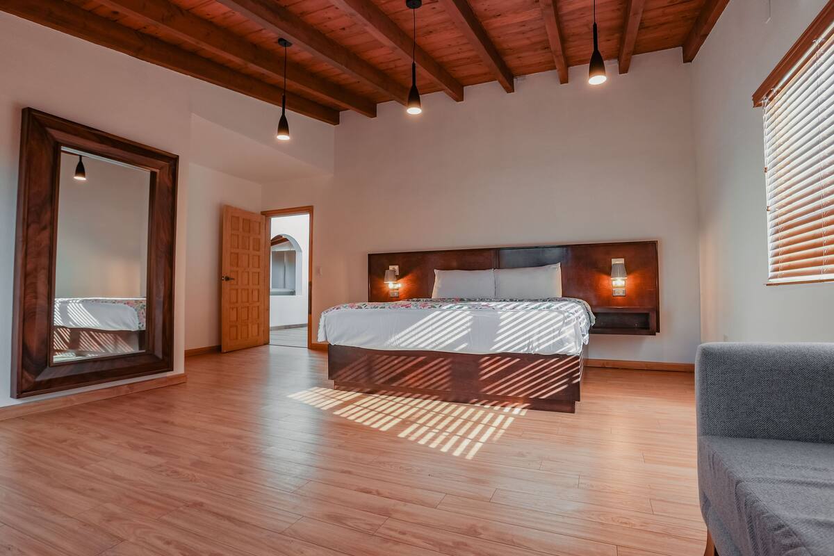 Ein Schlafzimmer mit Holzboden, einem großen Bett mit Kopfteil, zwei Nachttischlampen und einem großen Spiegel links.