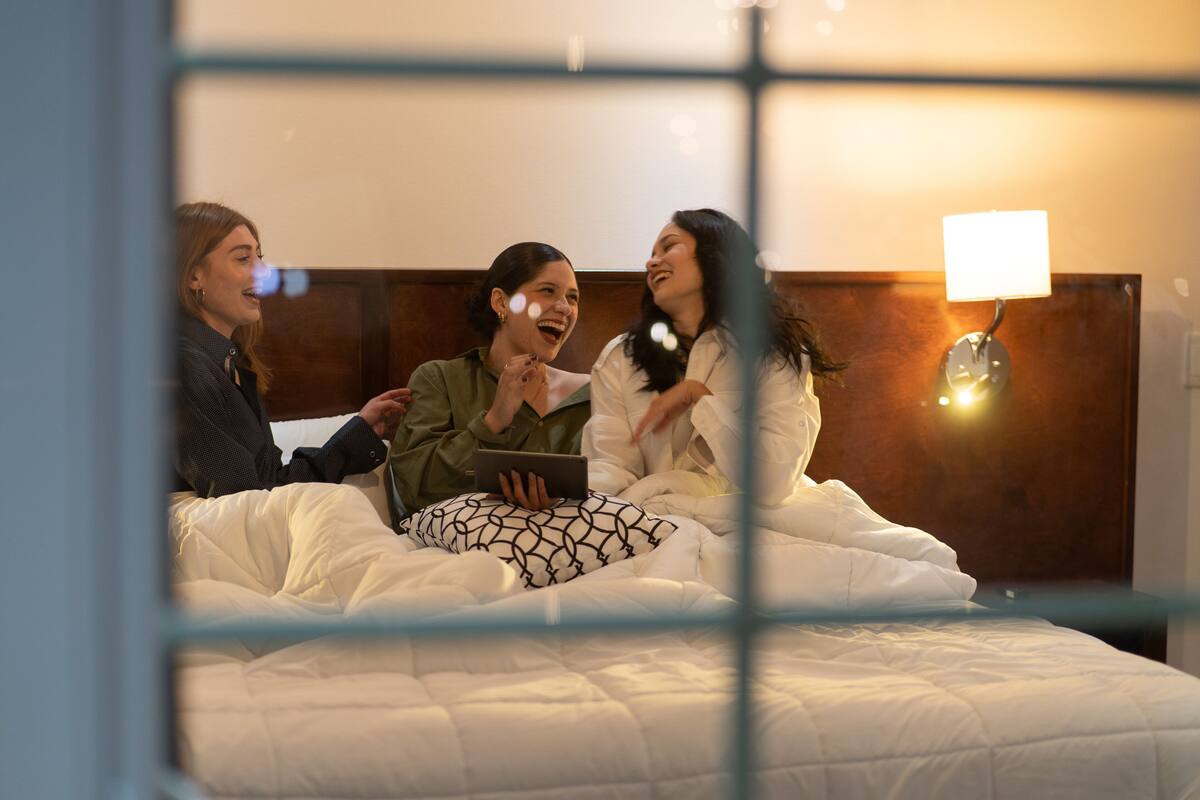Drei Personen sitzen auf einem Bett mit Kopfteil und Nachttischlampe.