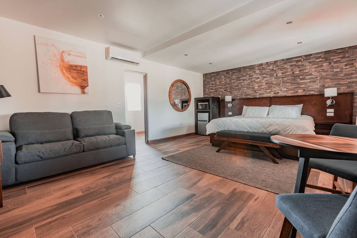 Ein modernes Hotelzimmer mit Holzboden, einem grauen Sofa, einem Bett mit Holz Kopfteil und einem kleinen Tisch.