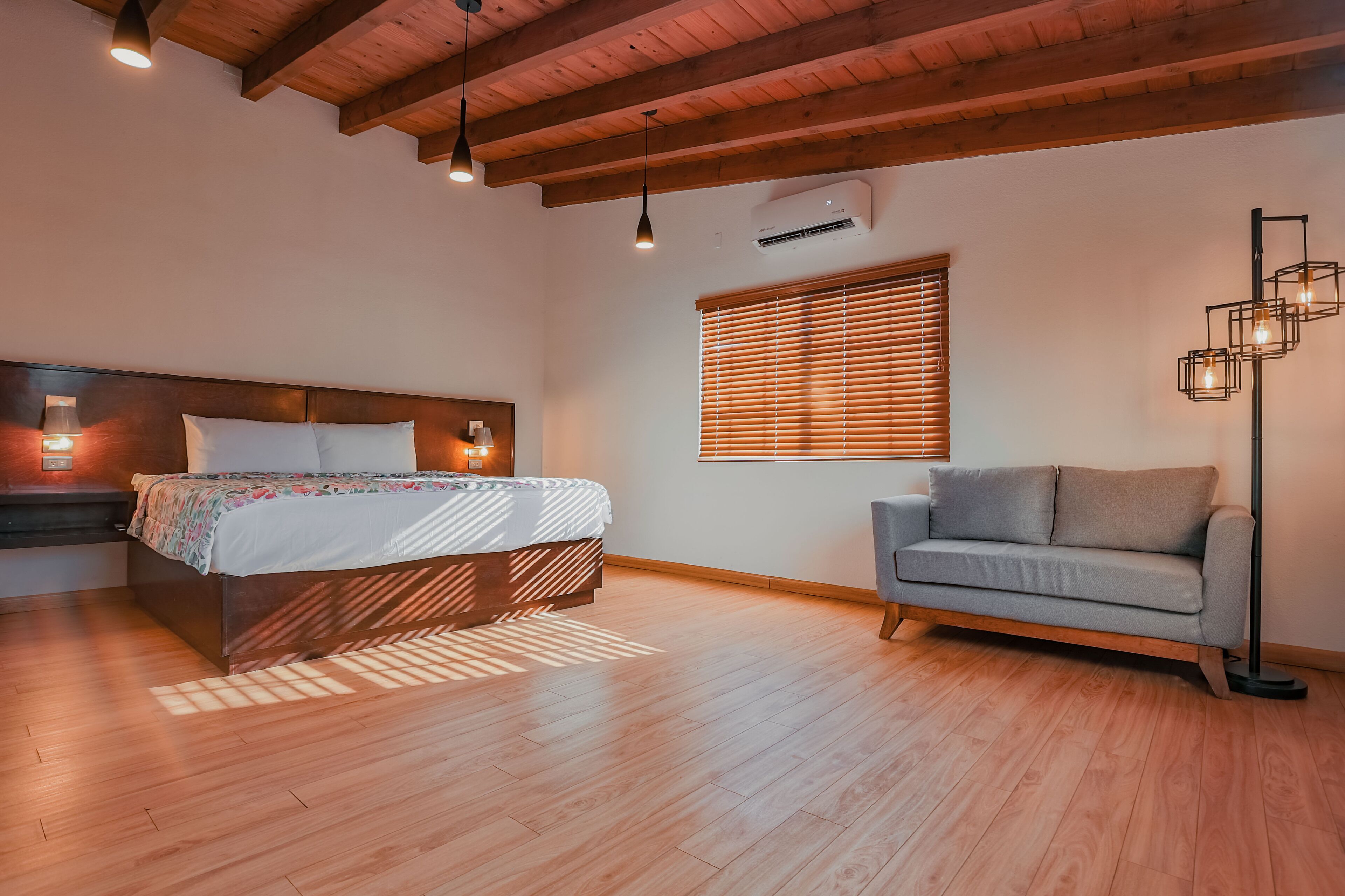 Un dormitorio con techo de madera, una cama con colcha estampada, un sofá gris y una lámpara de pie.