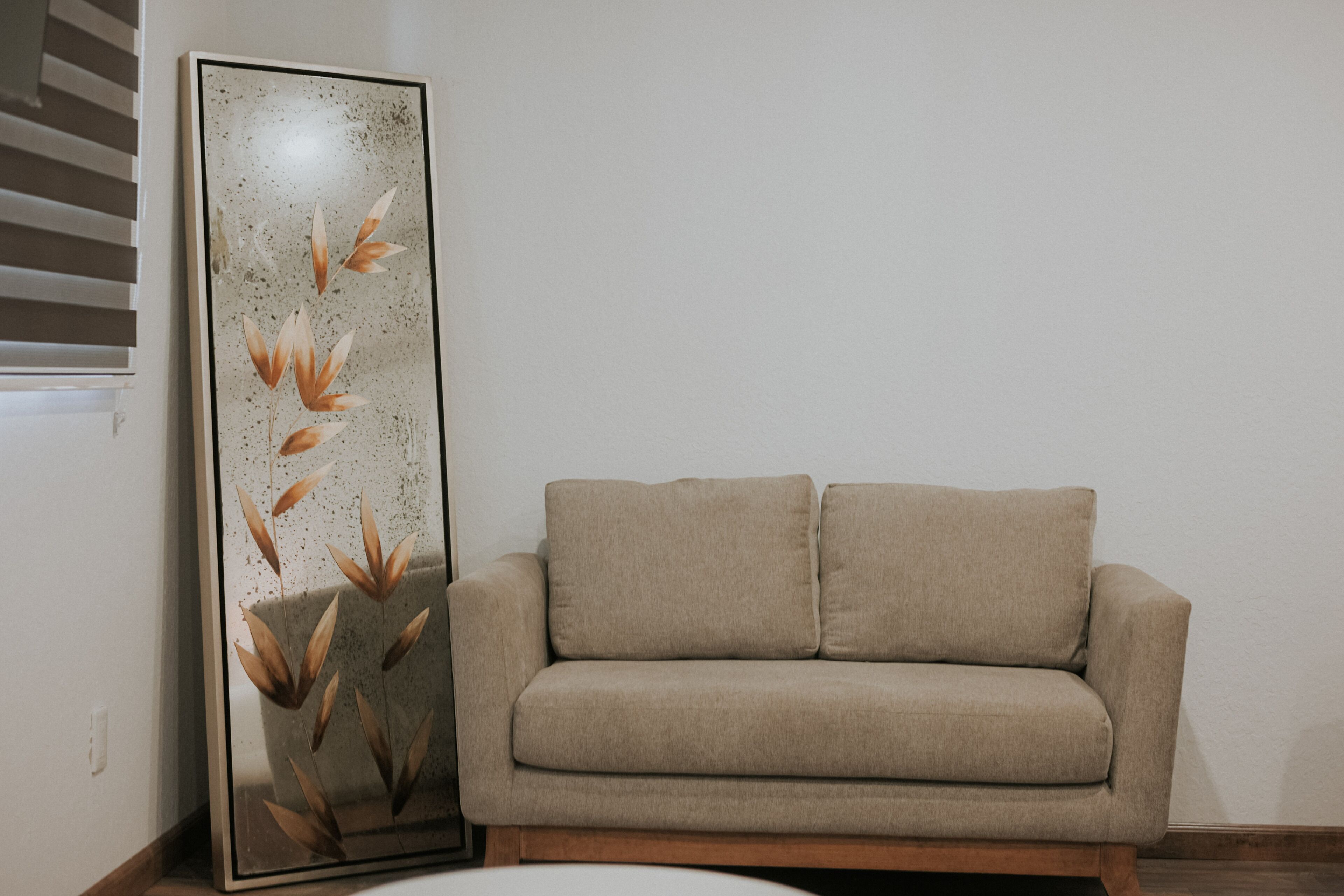 A beige sofa and a tall, narrow mirror with a floral design.