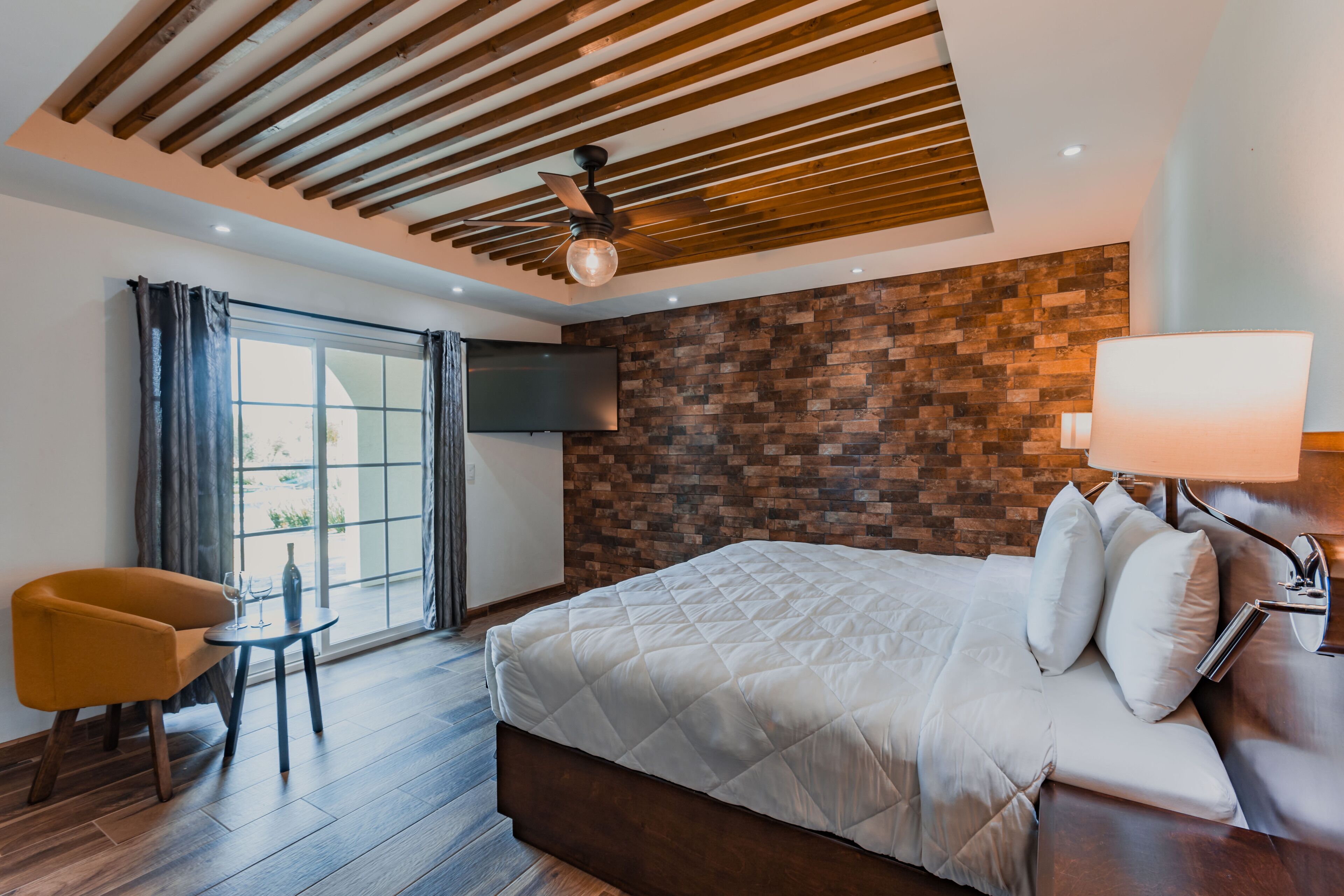 A modern hotel room with a large bed, a wooden ceiling, a brick wall, a TV, a small table, and a chair.
