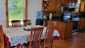 Un comedor con una mesa dispuesta para cuatro, sillas de madera, un mantel de encaje y vistas a través de una ventana con persianas.