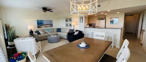 Un amplio salón con una mesa de madera para comer, sillas blancas, un sofá y una cocina al fondo.