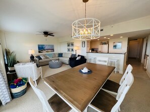 Un vaste espace de vie comprenant une table à manger en bois, des chaises blanches, un canapé et, au fond, une cuisine.
