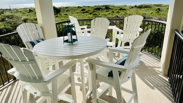 A white outdoor seating area with a round table and chairs, overlooking a beach and ocean.