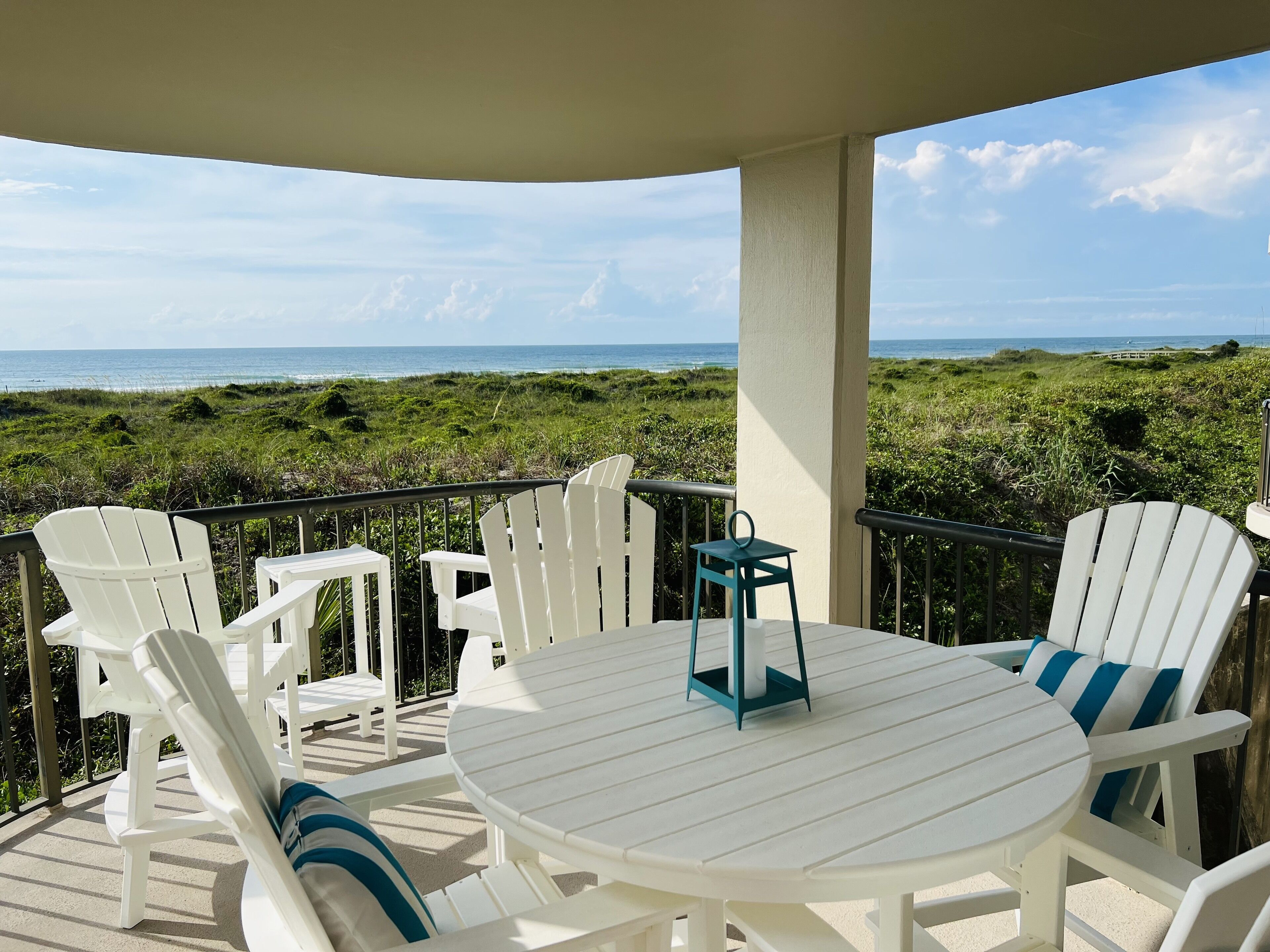 Un área de estar al aire libre cubierta con sillas blancas y una mesa redonda, con vistas a una playa y un paisaje cubierto de césped.