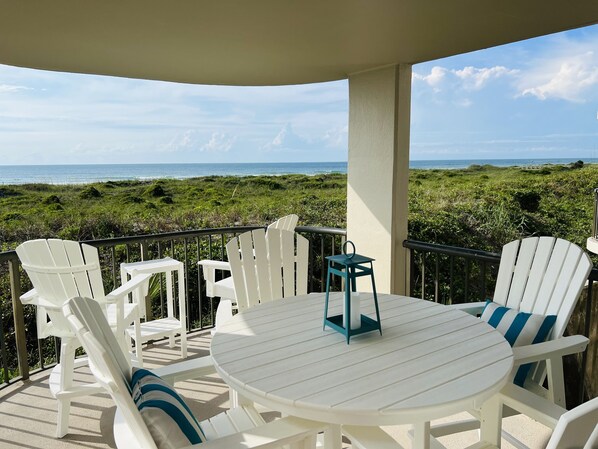Un espace extérieur aménagé avec des sièges, protégé par un toit, comprenant des chaises blanches et une table ronde, offrant une vue sur une plage et un paysage verdoyant.
