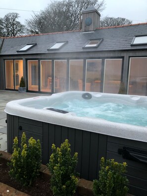 A hot tub with a control panel, surrounded by small potted plants, and a house with large sliding glass doors and a clock on the roof. - Modern Converted Stables in the heart of Pembrokeshire (Wiston)