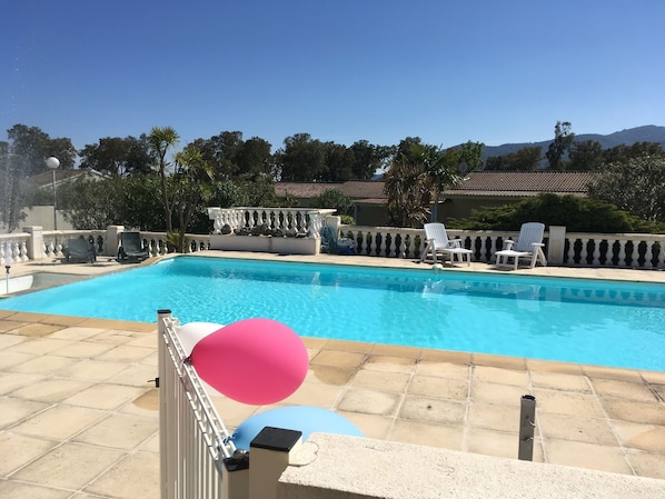 A swimming pool with tiled deck and white balustrades, surrounded by greenery and a building with a tiled roof.