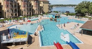 A multi-level pool area with various water features, including slides and a water play area, surrounded by lounge chairs and a view of a lake. - Top of Stillwater by Okoboji Luxury Rentals (Arnolds Park)