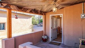 Un espacio exterior techado con techo de madera, un banco de hormigón, una planta en maceta y una puerta que da a un espacio interior.