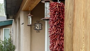 Un porche de madera con una decoración de chile colgante, un comedero para pájaros y un letrero con el número de la casa.