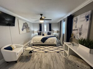 Un dormitorio con cama, televisión montada en la pared, ventilador de techo y un amplio ventanal con cortinas.