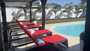 A covered outdoor area with a pool, red lounge chairs, and a white fence in the background. - ☀️WOW☀️ UPDATED with HEATED POOL & HOT TUB HOME 5 MIN TO BEACH 3BD 2BA 2 KING BEDS (Seminole)