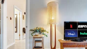 Una sala de estar moderna con un televisor de pantalla plana mostrando servicios de transmisión, una lámpara de pie y una silla de madera.