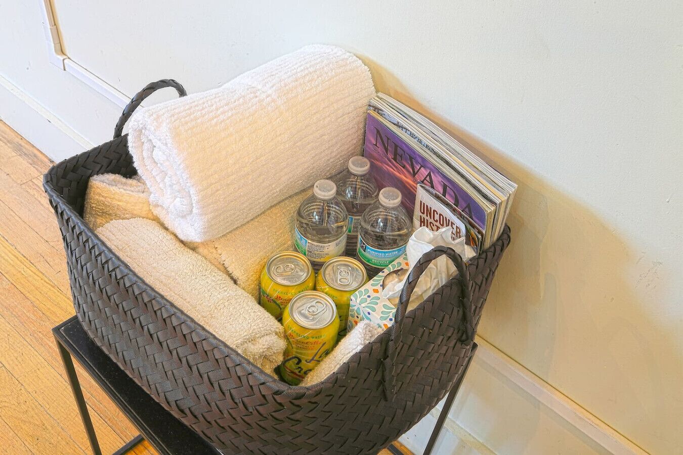 Una cesta tejida con una toalla blanca, varias latas de bebidas y una pila de revistas.