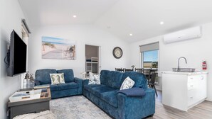 A modern living room with a blue sofa, a flat-screen TV, and a kitchen area with a white countertop and sink. - Hampton Cottage - " A Place To Call Home" (Port Albert)