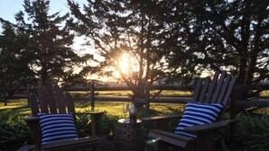 Two Adirondack chairs with striped cushions, a small table with a glass, and a view of a sunset behind trees. - Hampton Cottage - " A Place To Call Home" (Port Albert)
