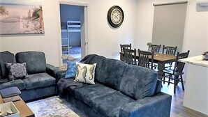 A living room with a grey sofa, a coffee table, and a dining area with a wooden table and chairs. - Hampton Cottage - " A Place To Call Home" (Port Albert)