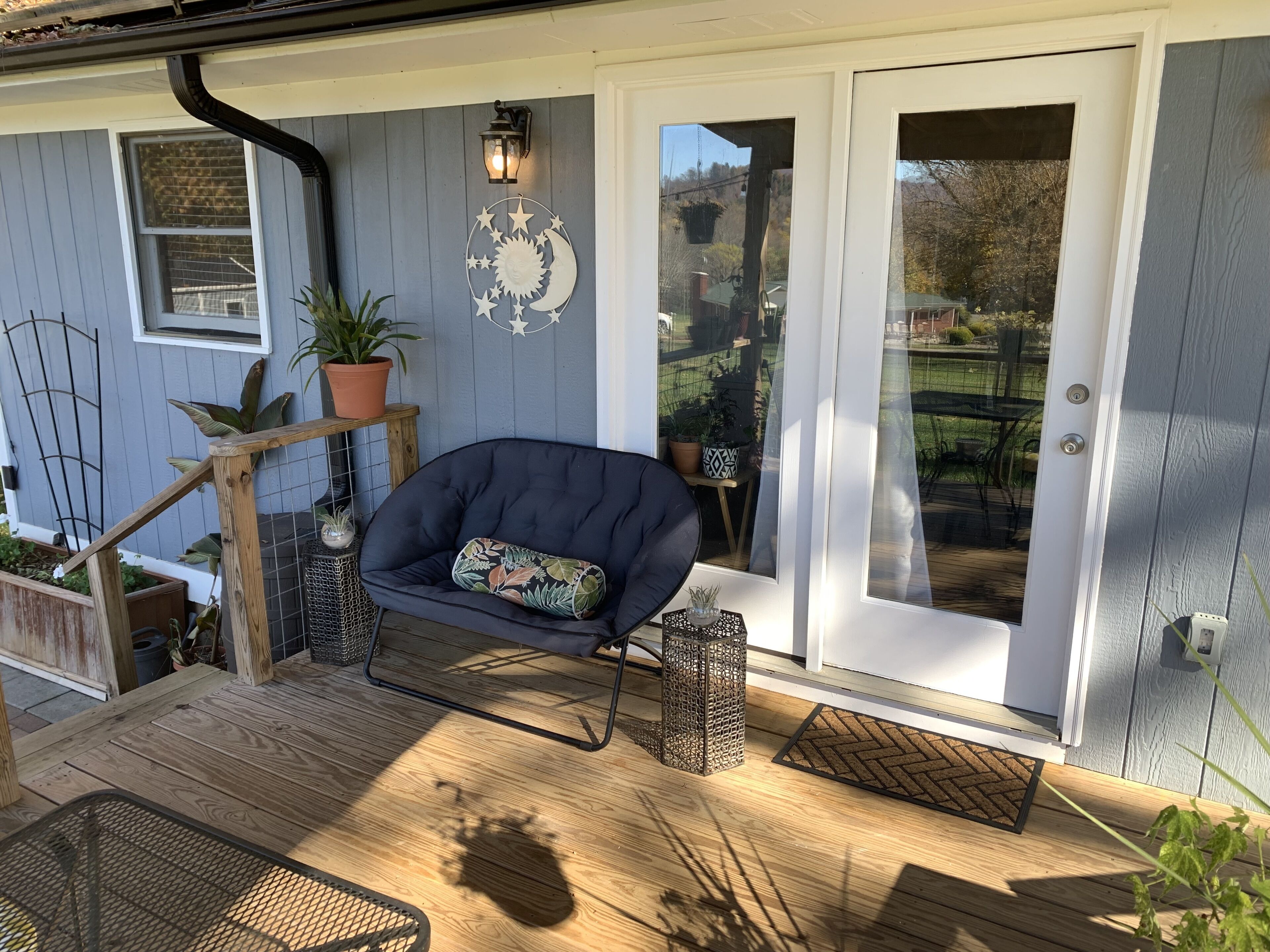 Een veranda met een zwarte stoel, een houten tafel, een bloempot, een metalen lantaarn en een decoratief wandobject.