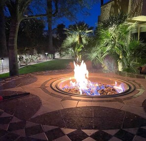 A circular outdoor fire pit surrounded by stone tiles, with a palm tree and other greenery in the background. - Desert Oasis in Sunrise Palms  (Palm Springs)
