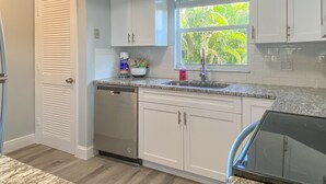 A modern kitchen with white cabinets, a built-in dishwasher, a granite countertop, and a window with a view of greenery. - Spacious Beach Retreat, Steps to Sugary Sand, Conveniences and Fun! (Fort Myers Beach)