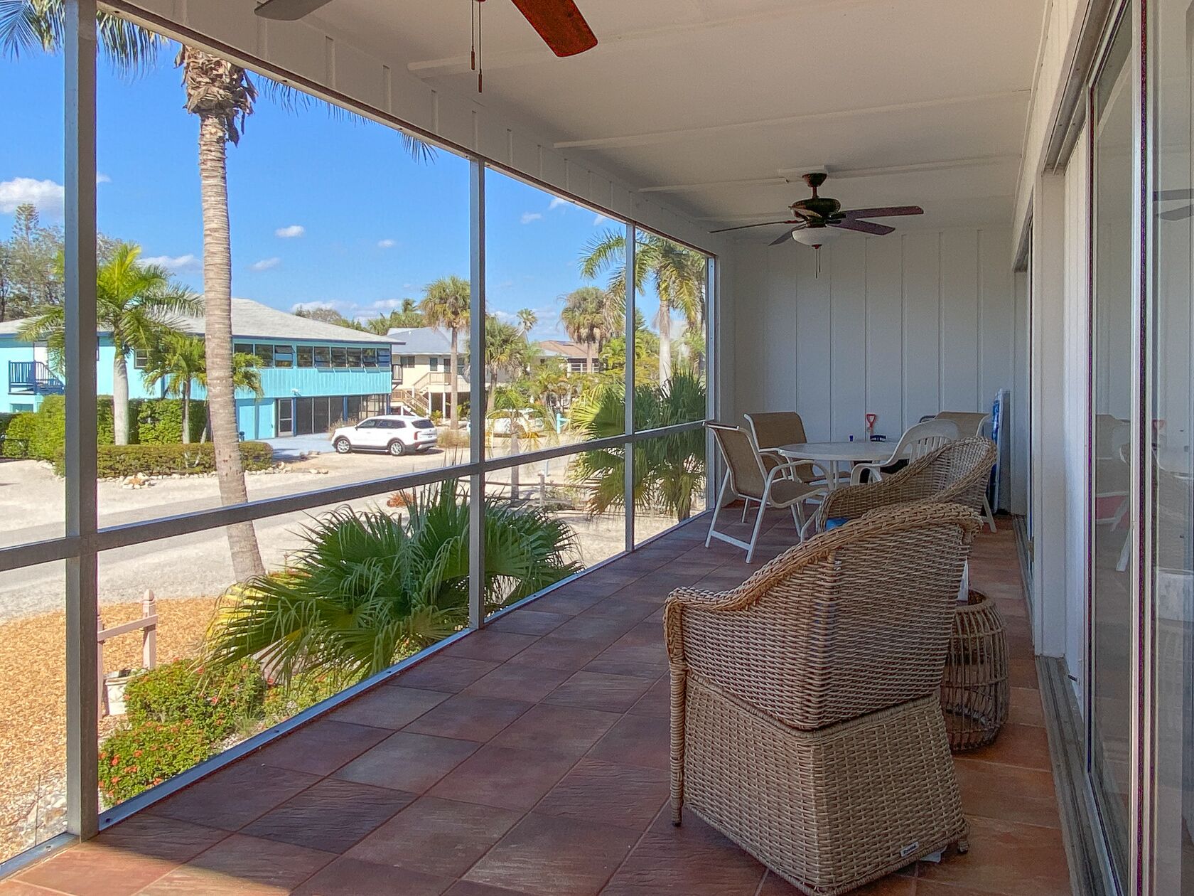 A covered outdoor area with wicker furniture, a ceiling fan, and a view of a street with palm trees and buildings.