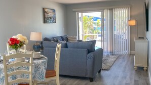 A living room with a grey sofa, a dining table with chairs, a painting on the wall, and a view through a window. - Spacious Beach Retreat, Steps to Sugary Sand, Conveniences and Fun! (Fort Myers Beach)