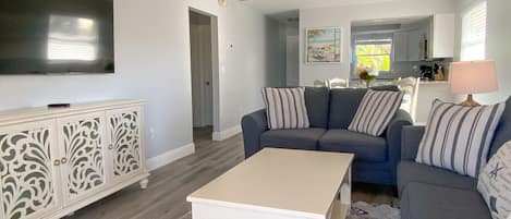 A living room with a sofa, a coffee table, and a TV. There is a kitchen in the background.