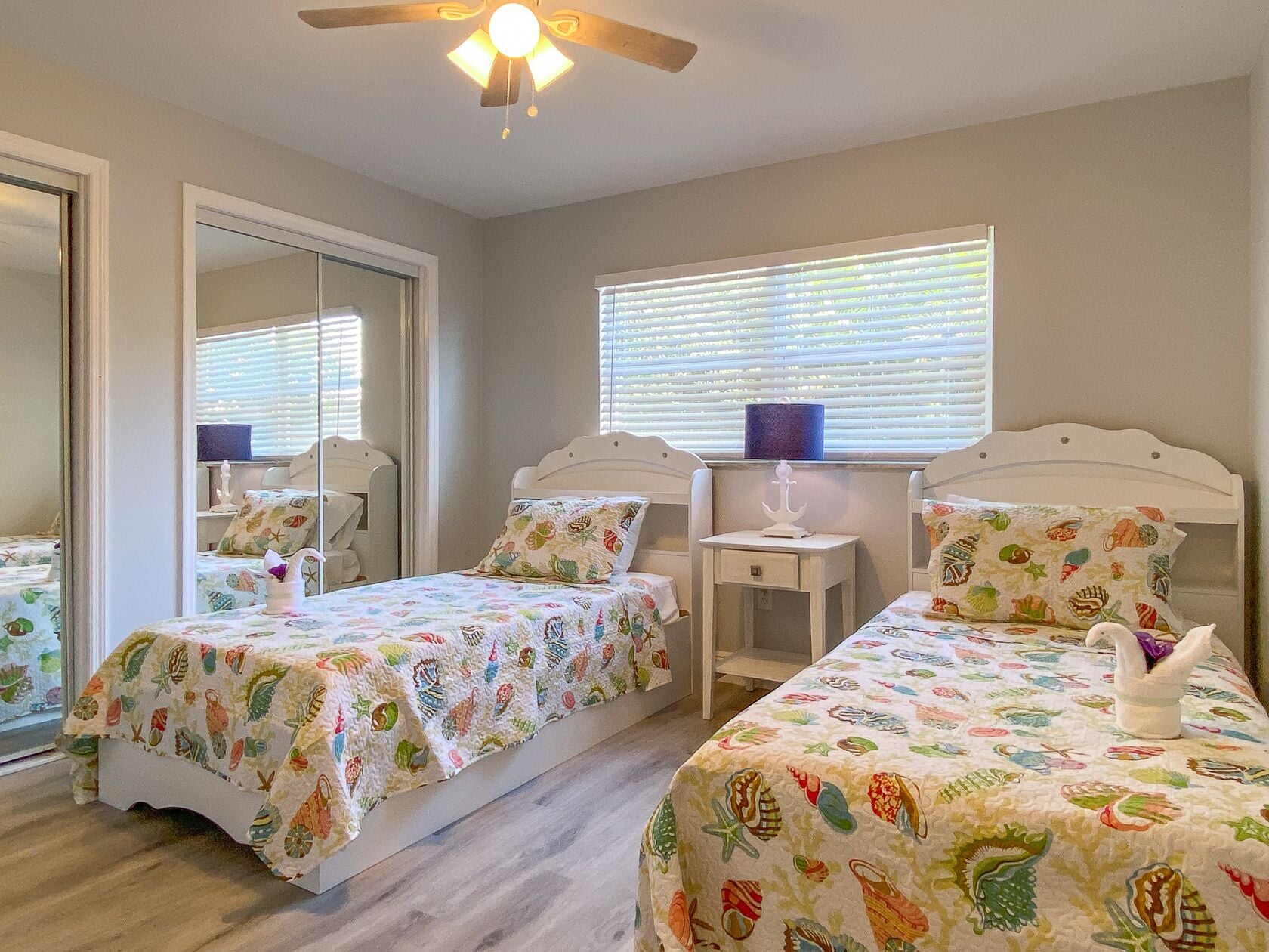 A bedroom with two single beds, a ceiling fan, a window with blinds, and a mirror.