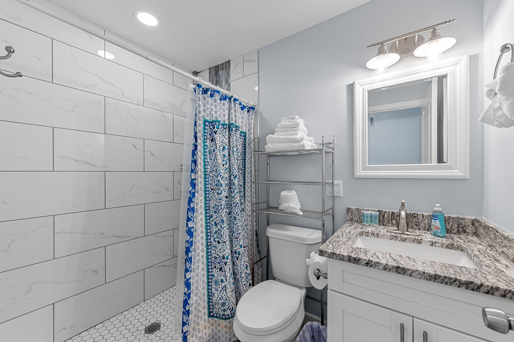Une salle de bain moderne avec des carreaux de marbre blanc, une douche avec un rideau à motifs bleus, des toilettes, un lavabo avec un plan de toilette en marbre et un miroir au-dessus du lavabo.
