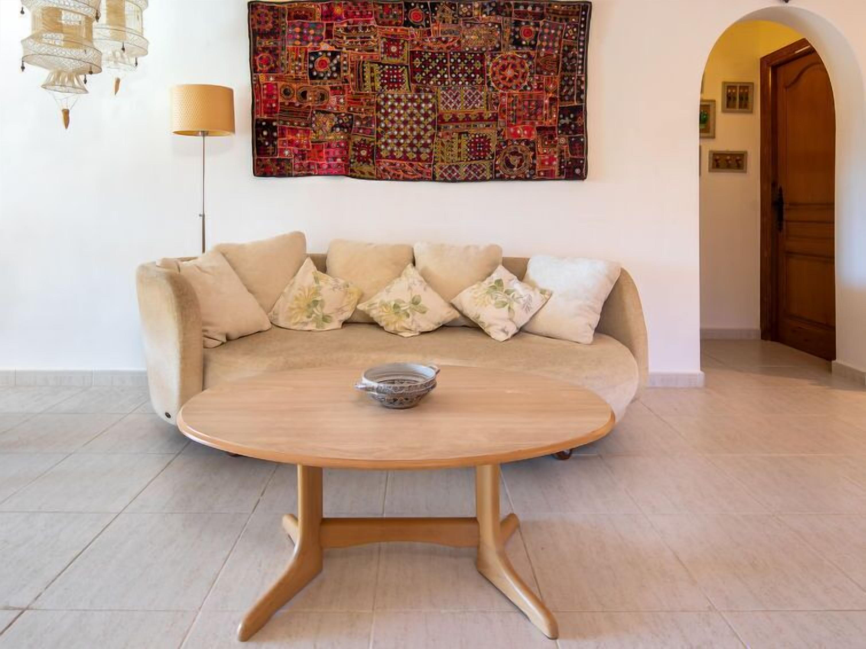 A modern living room with a beige sofa, a round wooden coffee table, and a colorful patchwork wall hanging.