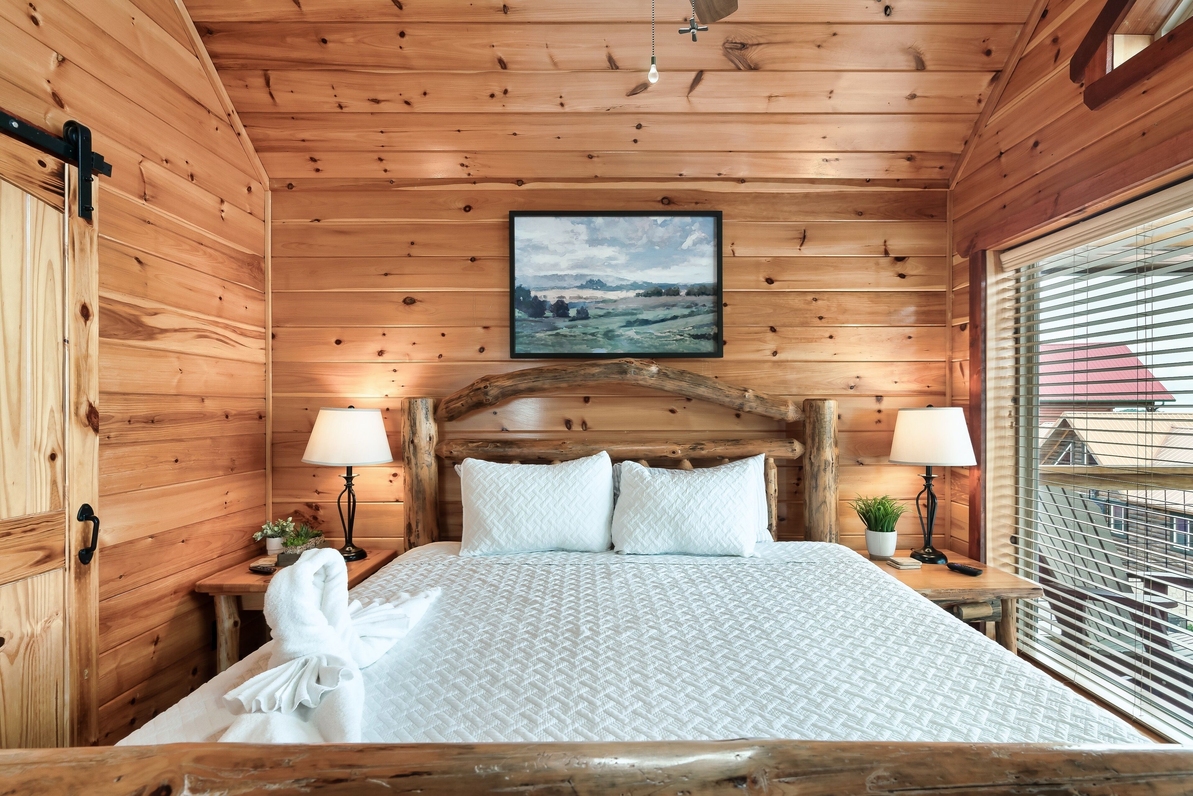 Ein schlichtes Schlafzimmer im Chalet-Stil mit einem Bett, zwei Nachttischlampen, einem gerahmten Bild an der Wand und einem Fenster mit Jalousien.