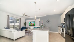 Uma cozinha moderna com uma geladeira grande, um espaço de jantar com uma mesa e cadeiras brancas, e uma sala de estar com um sofá branco e uma televisão.